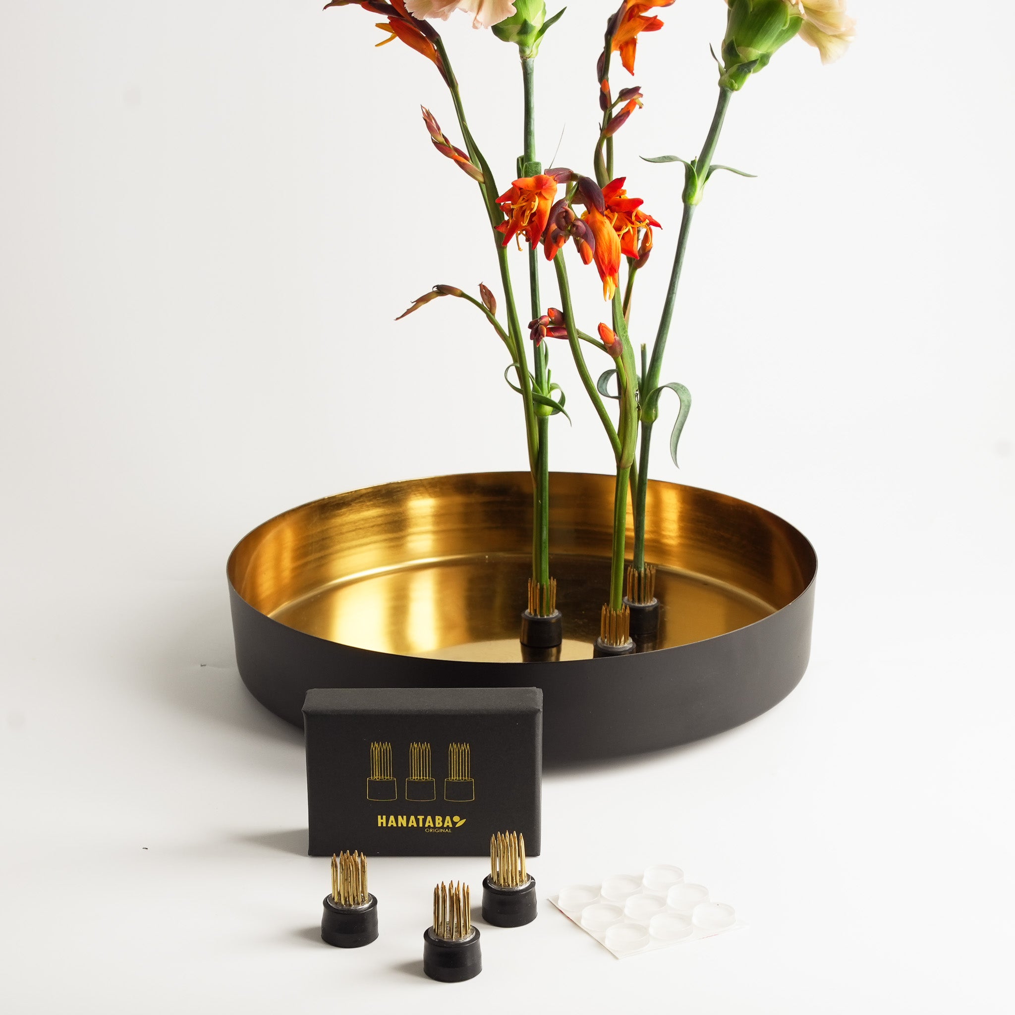 Three Hanataba magnetic flower frogs holding orange and pink flowers upright inside a decorative gold-lined bowl. The magnets and silicone pads keep the arrangement stable.