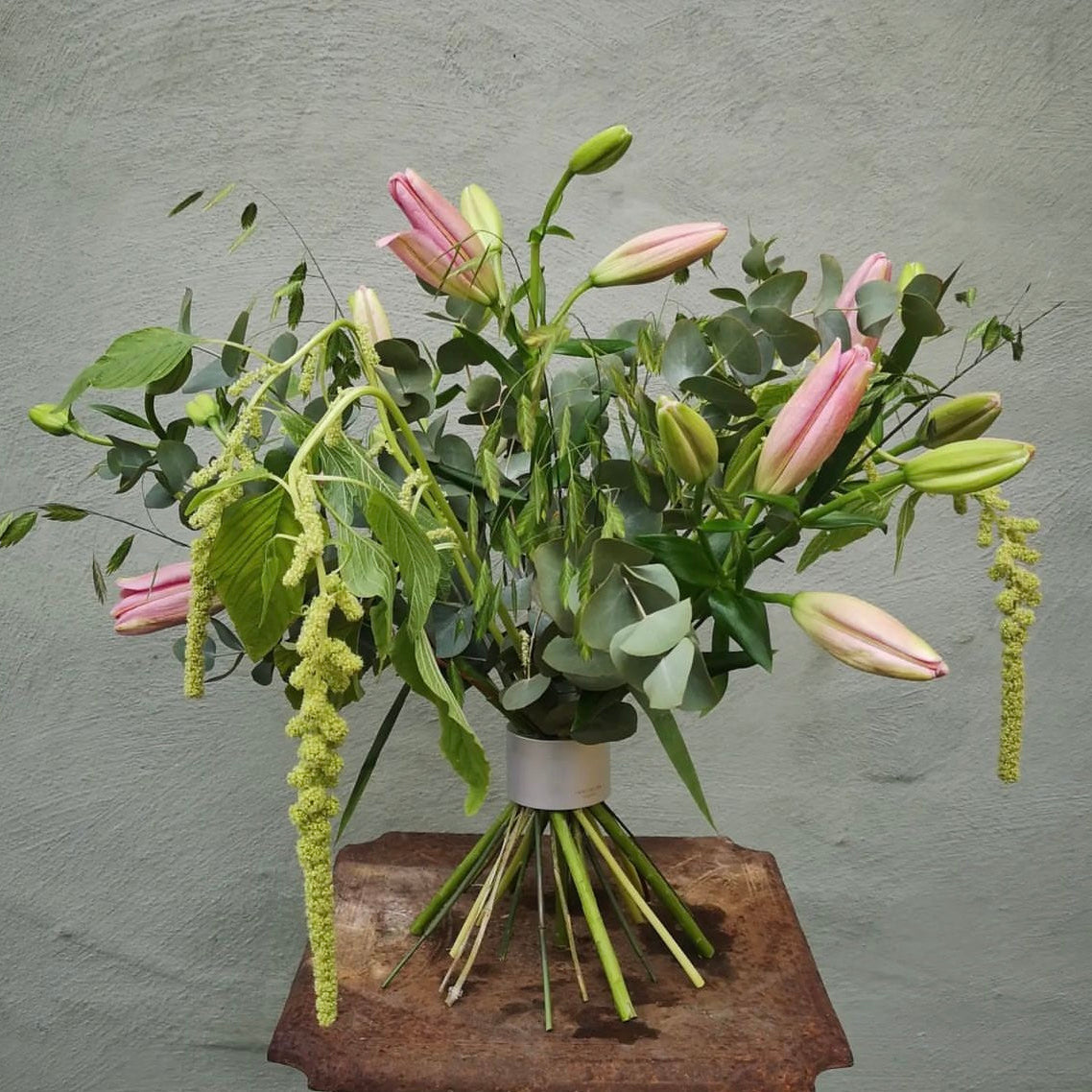 A lush floral arrangement featuring pink lilies and cascading green accents, all held together by a Hanataba Bouquet Twister in pearly silver, against a textured grey wall