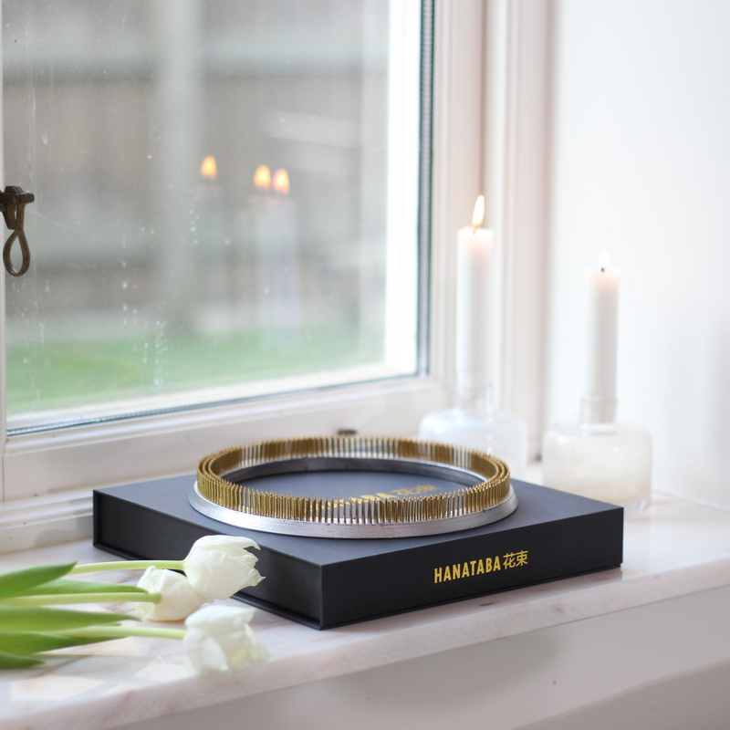 A Hanataba branded black box with a golden kenzan ring on a windowsill, accompanied by white tulips and lit candles, creating a serene ambiance.
