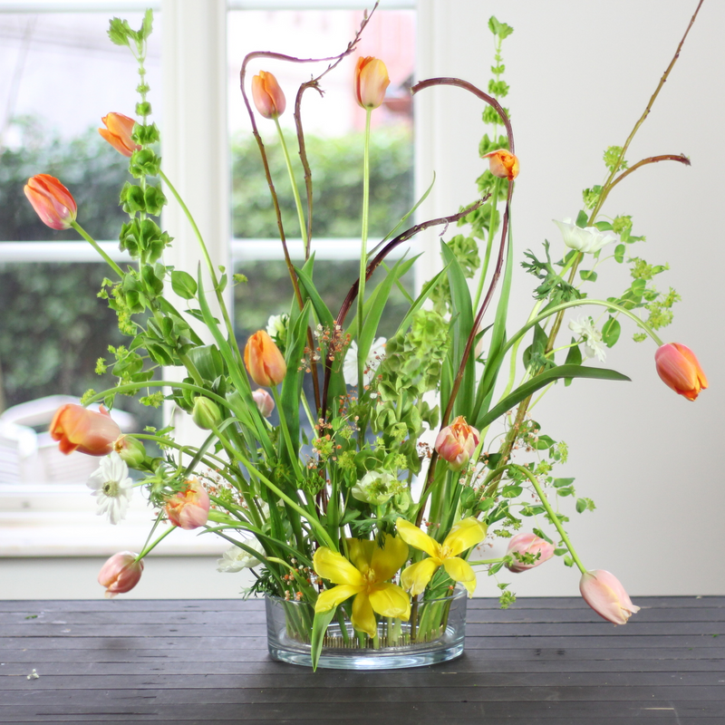 A vibrant arrangement of tulips and mixed greenery standing upright in a 200mm kenzan ring, placed on a dark wooden table near a window.
