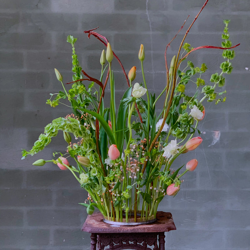 A vibrant bouquet featuring tulips and an array of greenery, meticulously arranged with a 200mm Kenzan ring on an ornate wooden pedestal against a grey brick wall.