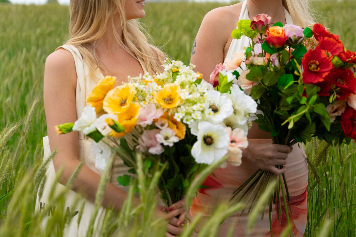 Two women standing in a grassy field, each holding a large, vibrant bouquet of artificial flowers. The woman on the left has blonde hair and wears a cream sleeveless top, while the woman on the right wears glasses and a white halter top.