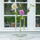 Three floral candle vase holders with Dahlias and green flowers on a white table in front of a window.
