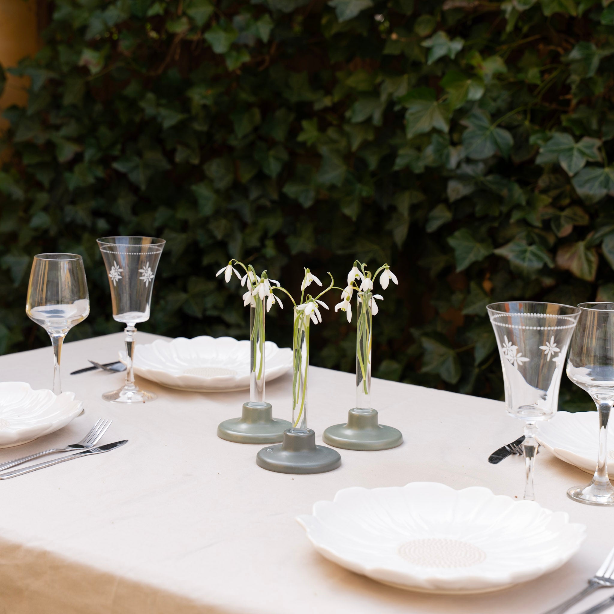 Dining table set with white plates, glasses, and small floral arrangements in a set of three candle vases against a green leafy background.