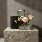 A black rectangular gift box with gold lettering sits beside a clear round vase filled with mixed flowers, displayed on a rough stone pedestal against a textured gray wall.