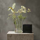 A clear glass vase filled with white and pale yellow dried flowers sits on a light stone pedestal next to a black rectangular box with a gold oval logo, set against a textured gray concrete wall.