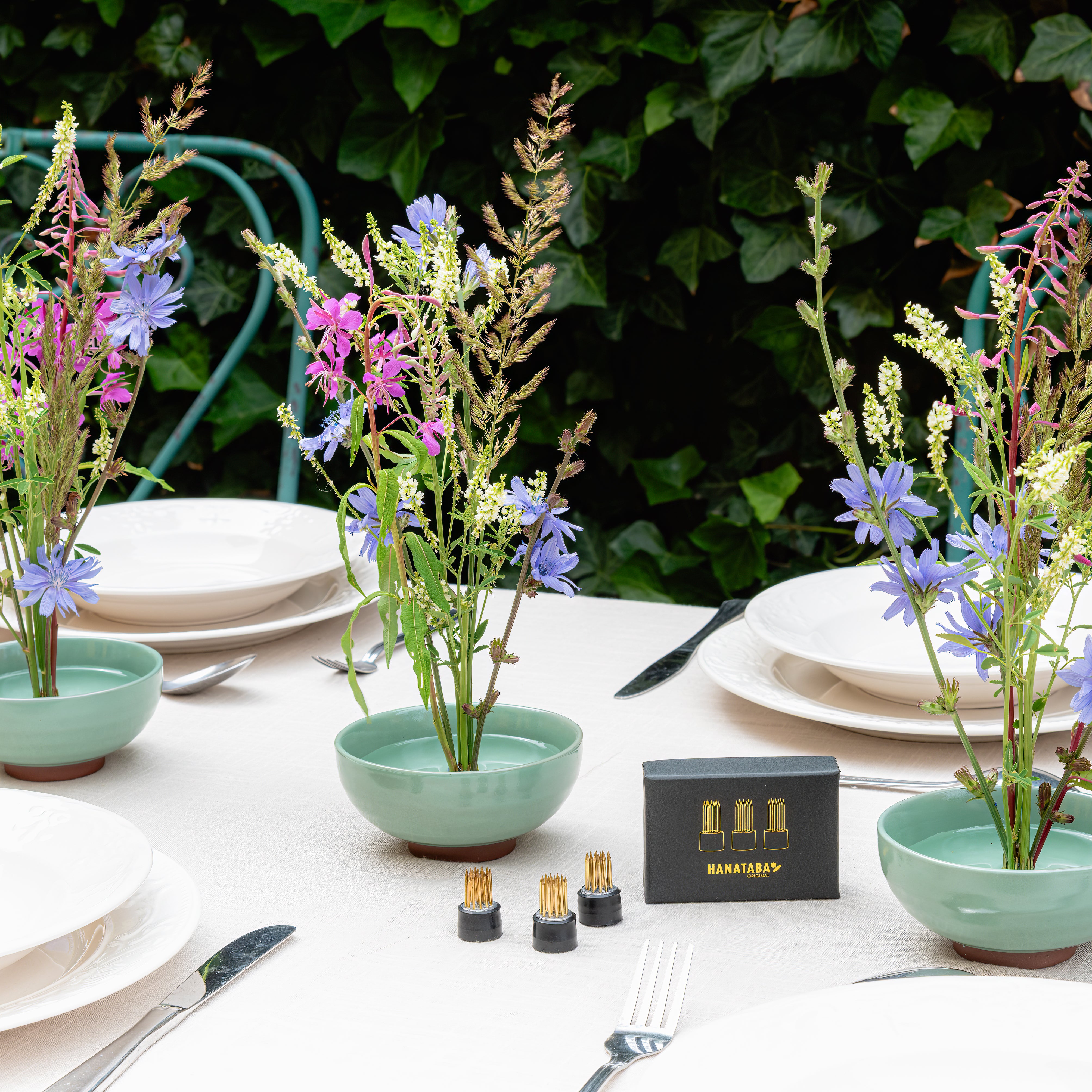 Table setting with floral arrangements in turquoise ceramic kenzan bowls, standing cutlery against a green leafy background.