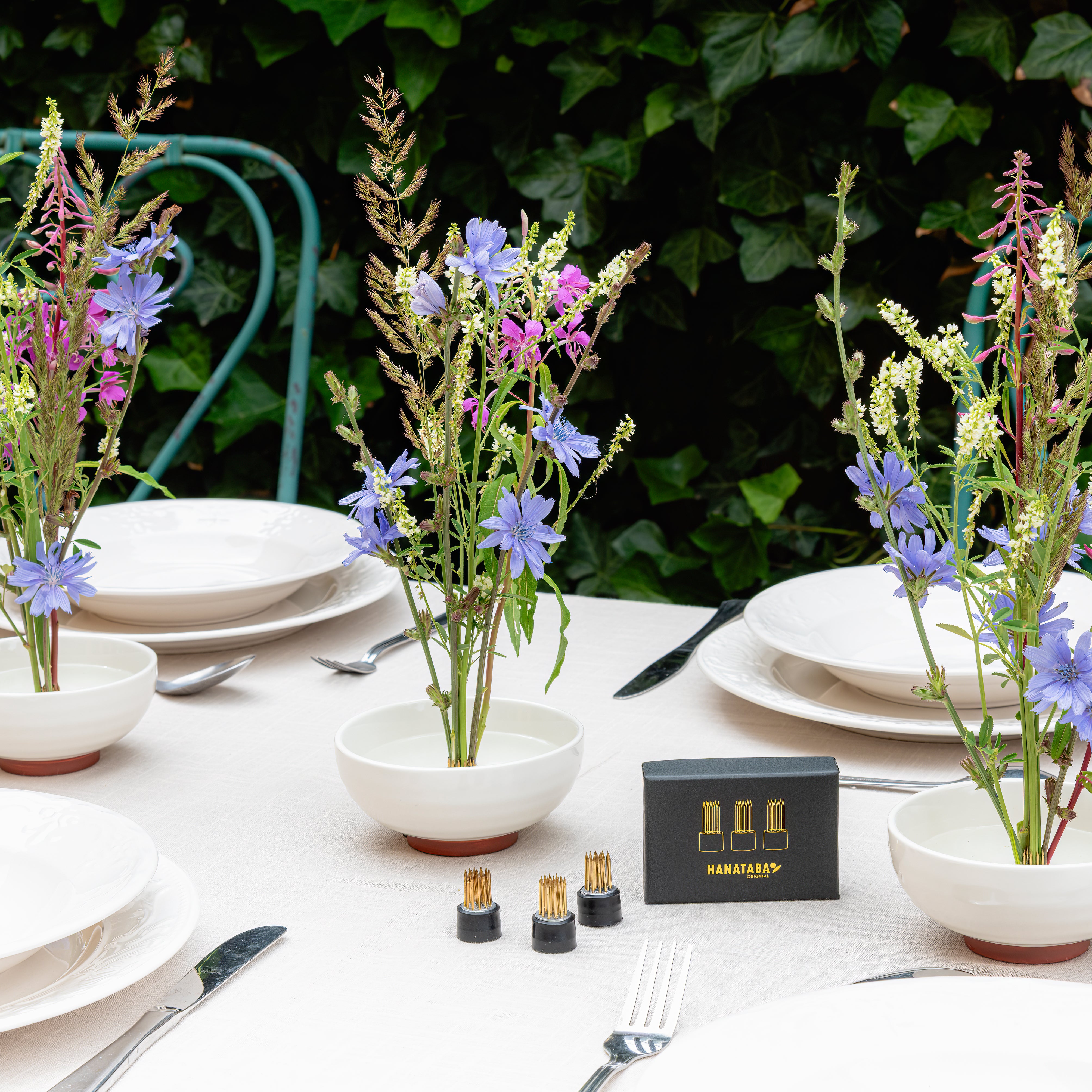 Table setting with floral arrangements in white ceramic bowls, plates, and cutlery against a green outdoor background.