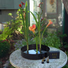 A black circular water-based flower arrangement metal plate with three tall green stems and orange calla lilies stuck to three Hanataba Magnetic MiniKenzan, displayed on a round stone table outdoors with a blurred garden background. A small black box labeled 'HANATABA' and three small magnetic mini kenzan sit beside the holder.