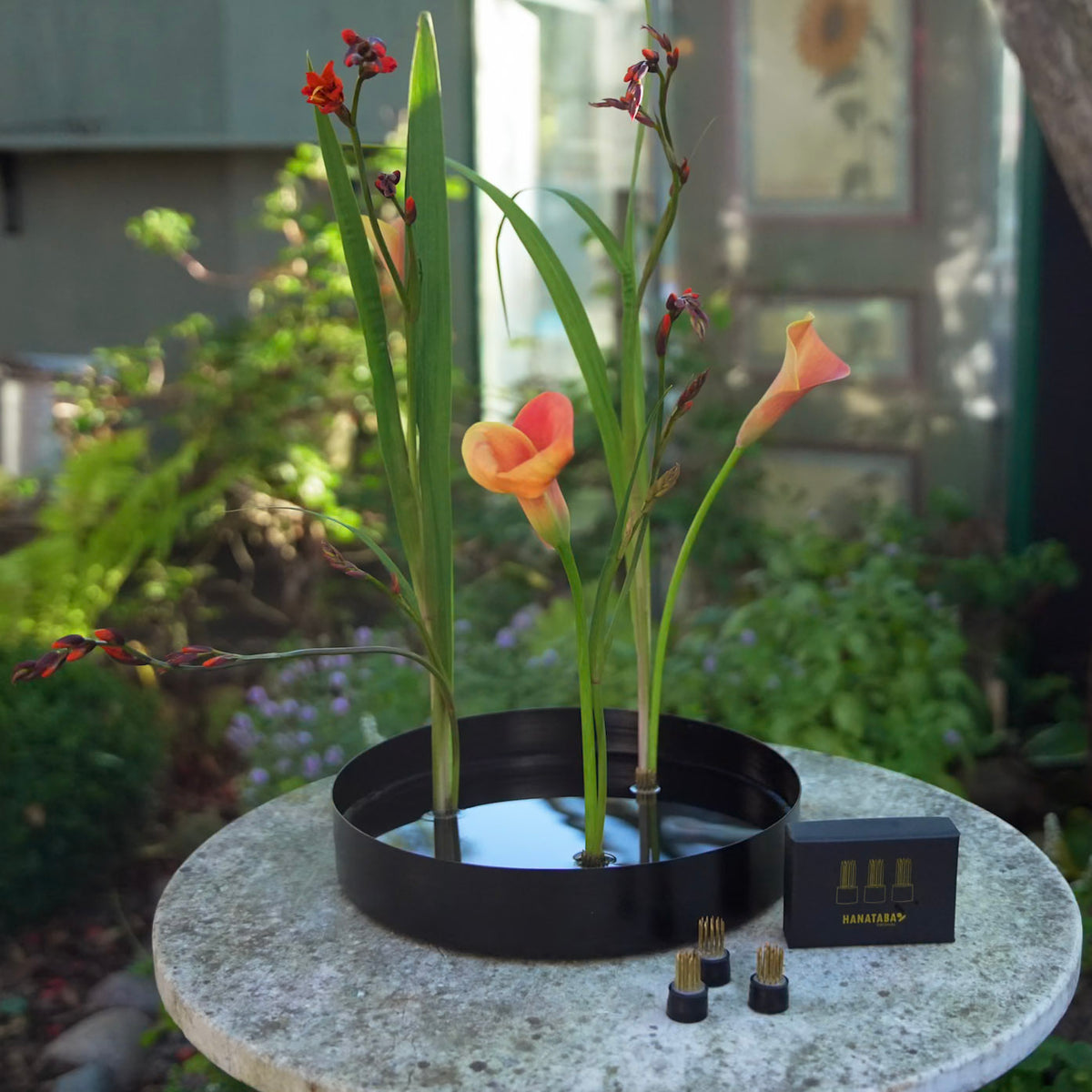 A black circular water-based flower arrangement metal plate with three tall green stems and orange calla lilies stuck to three Hanataba Magnetic MiniKenzan, displayed on a round stone table outdoors with a blurred garden background. A small black box labeled 'HANATABA' and three small magnetic mini kenzan sit beside the holder.