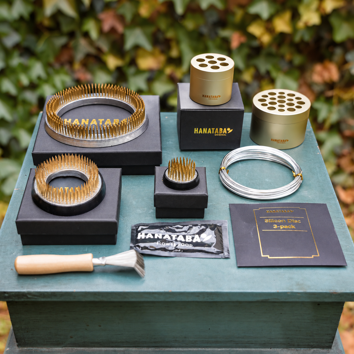 A set of Hanataba flower arrangement tools and accessories arranged on a painted wooden table outdoors, with blurred green and yellow foliage in the background. The set includes gold and silver metal kenzan rings, 70 mm and 120 mm ring, a silver bonsai wire, a small kenzan rake with a wooden handle, flower nutrition pouches, and hanataba champagne gold bouquet flower twisters.