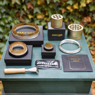A set of Hanataba flower arrangement tools and accessories arranged on a painted wooden table outdoors, with blurred green and yellow foliage in the background. The set includes gold and silver metal kenzan rings, 70 mm and 120 mm ring, a silver bonsai wire, a small kenzan rake with a wooden handle, flower nutrition pouches, and hanataba champagne gold bouquet flower twisters.