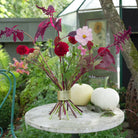 A floral arrangement in a gold-toned Hanataba Bouquet Twister placed on a stone table in a garden greenhouse setting, with two white pumpkins beside it.