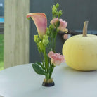 A small floral arrangement in a kenzan flower frog pin holder 34 mm, placed on a white table next to a pale yellow pumpkin. The bouquet includes a pink-and-cream calla lily, pink flowers, green buds, and green leaves.