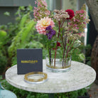 A clear glass vase with a kenzan ring 120 mm, filled with a colorful floral arrangement, sits on a round stone table outdoors. A black Hanataba Original box and a flower frog kenzan ring 120 mm are placed beside it, with green garden foliage in the background.