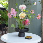 A black floral stand holding pink and cream dahlias next to a Hanataba Original Kenzan ring 70 mm, set on a white round table outdoors with a green fence in the background.