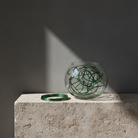 Glass vase with green abstract hanataba bonsai wire used for floral arrangements design, standing on a stone surface with a dark background