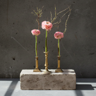 Three small hanataba candle vases  with pink ranunculus flowers on a concrete block against a gray wall.