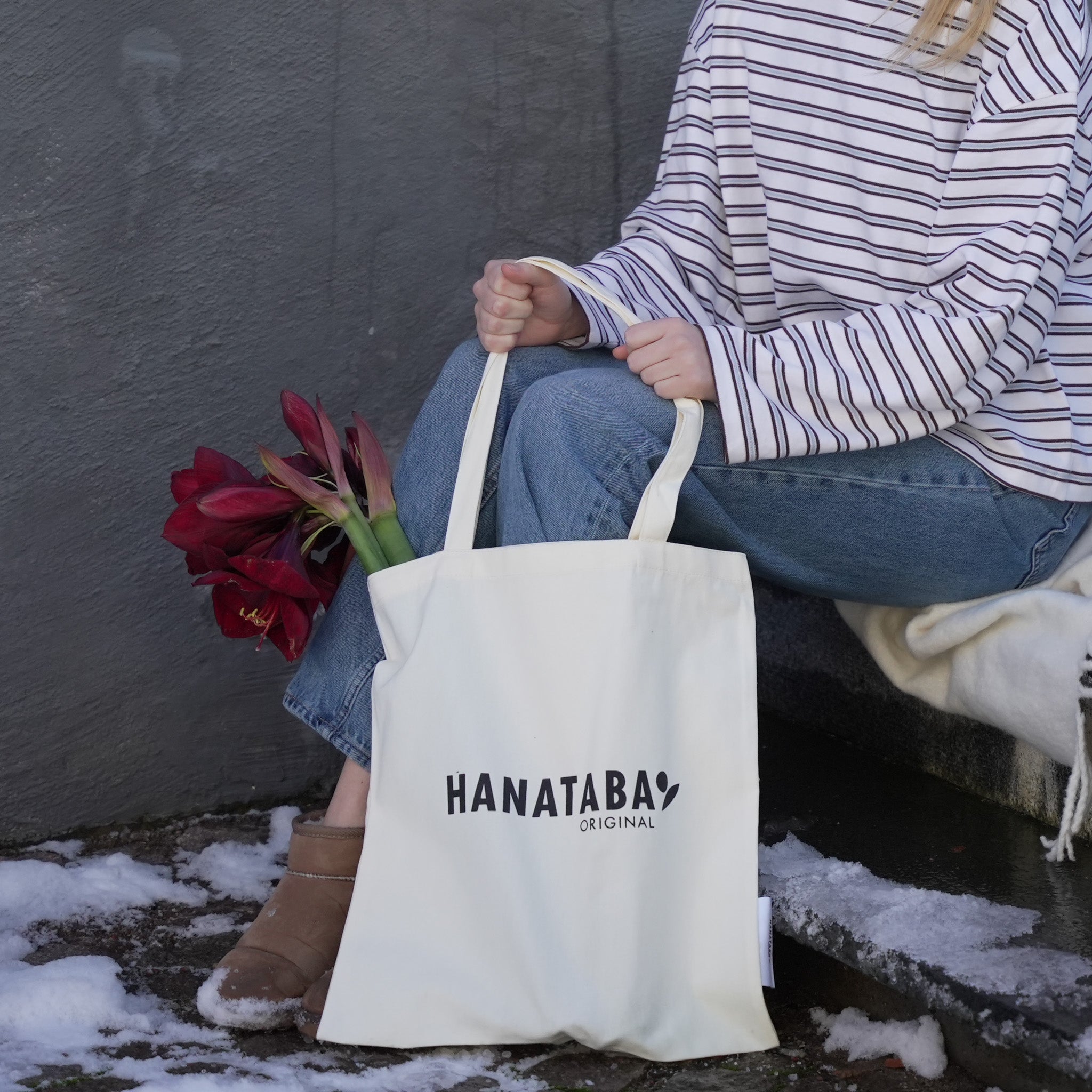 A person sitting on snowy steps holding a white Hanataba Original tote bag with red amaryllis flowers placed beside it.