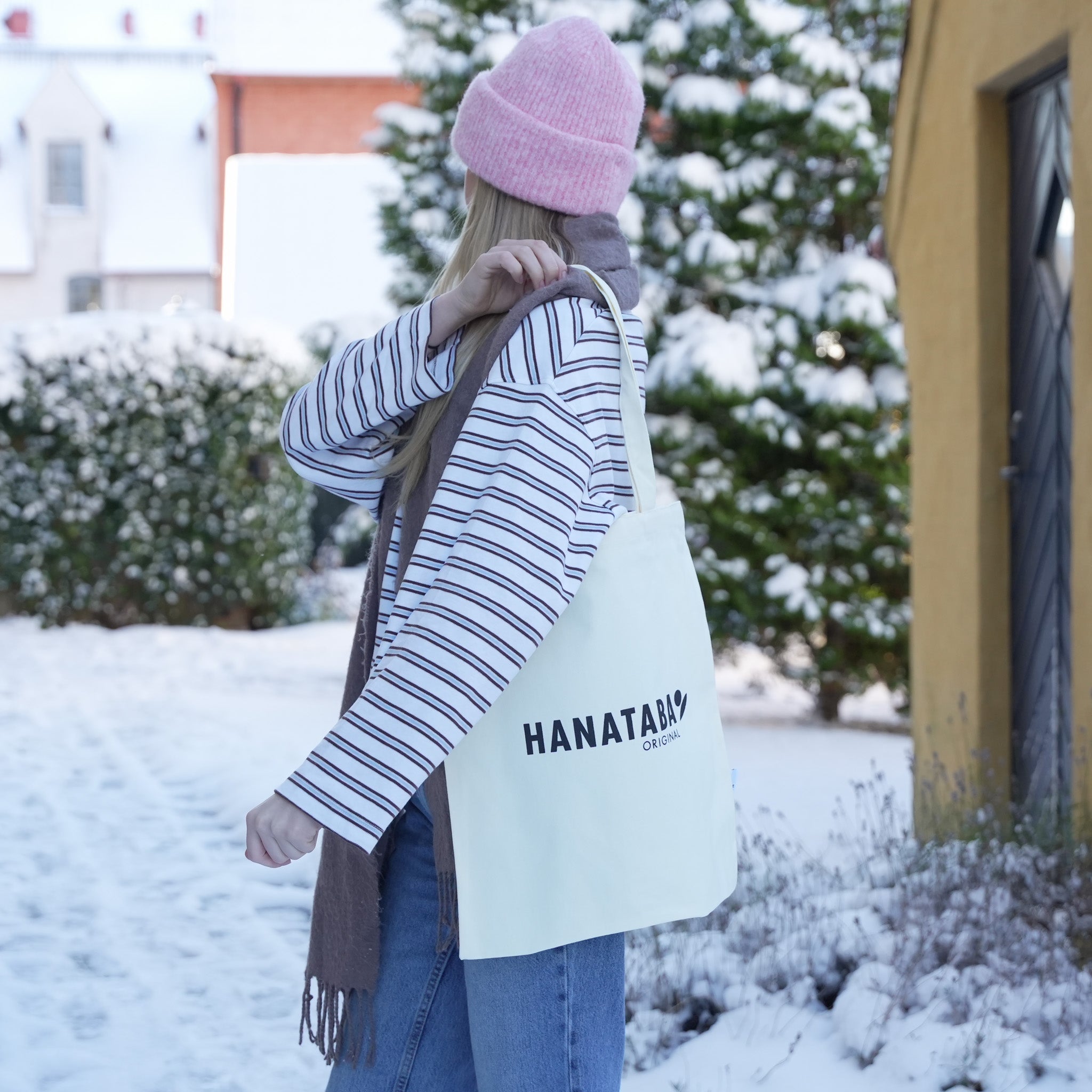 A woman wearing a pink beanie carries a white Hanataba Original tote bag while walking through a snowy outdoor scene.