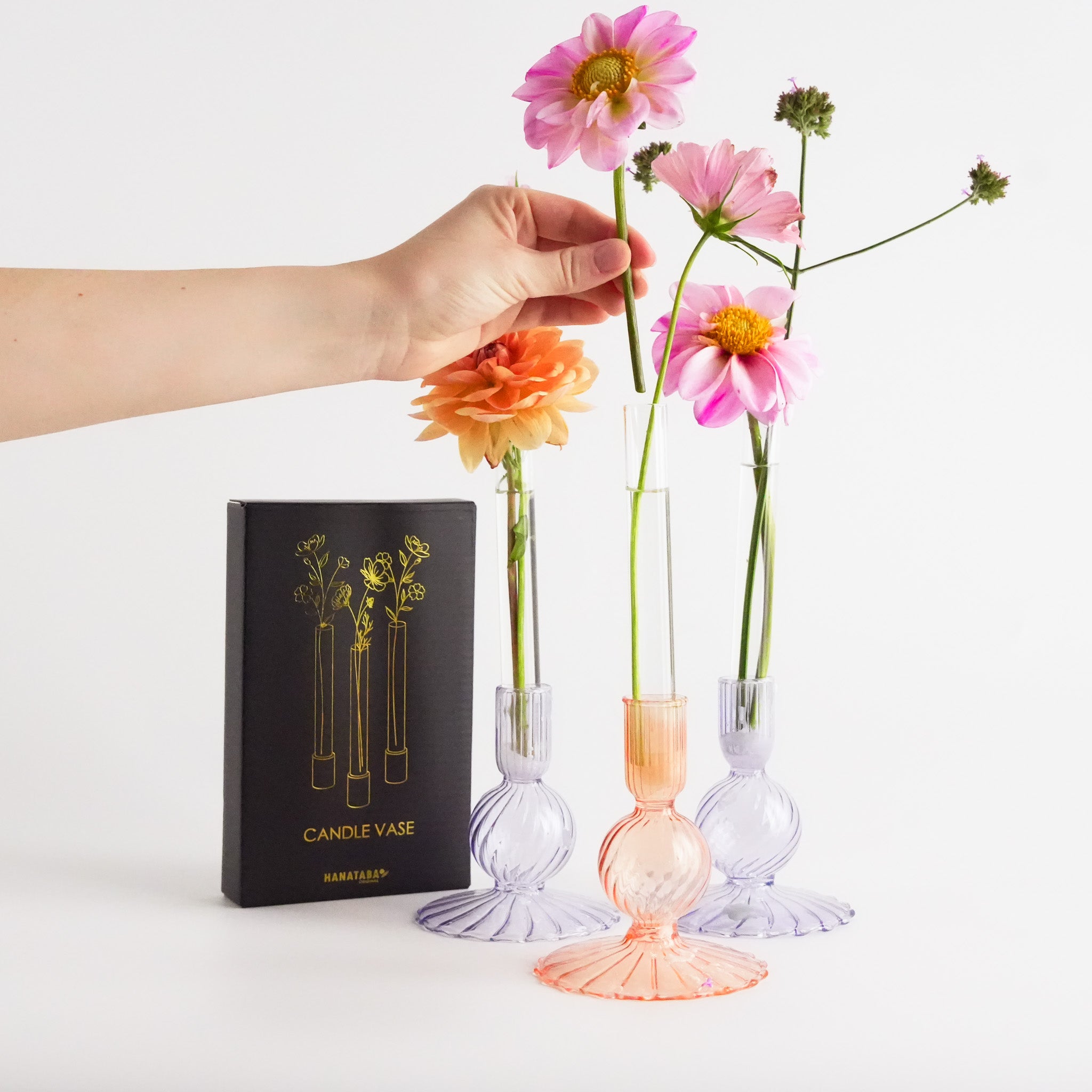 Hand holding a pink flower above three glass vases with flowers, one of which is the 'Candle Vase' by Hanataba.