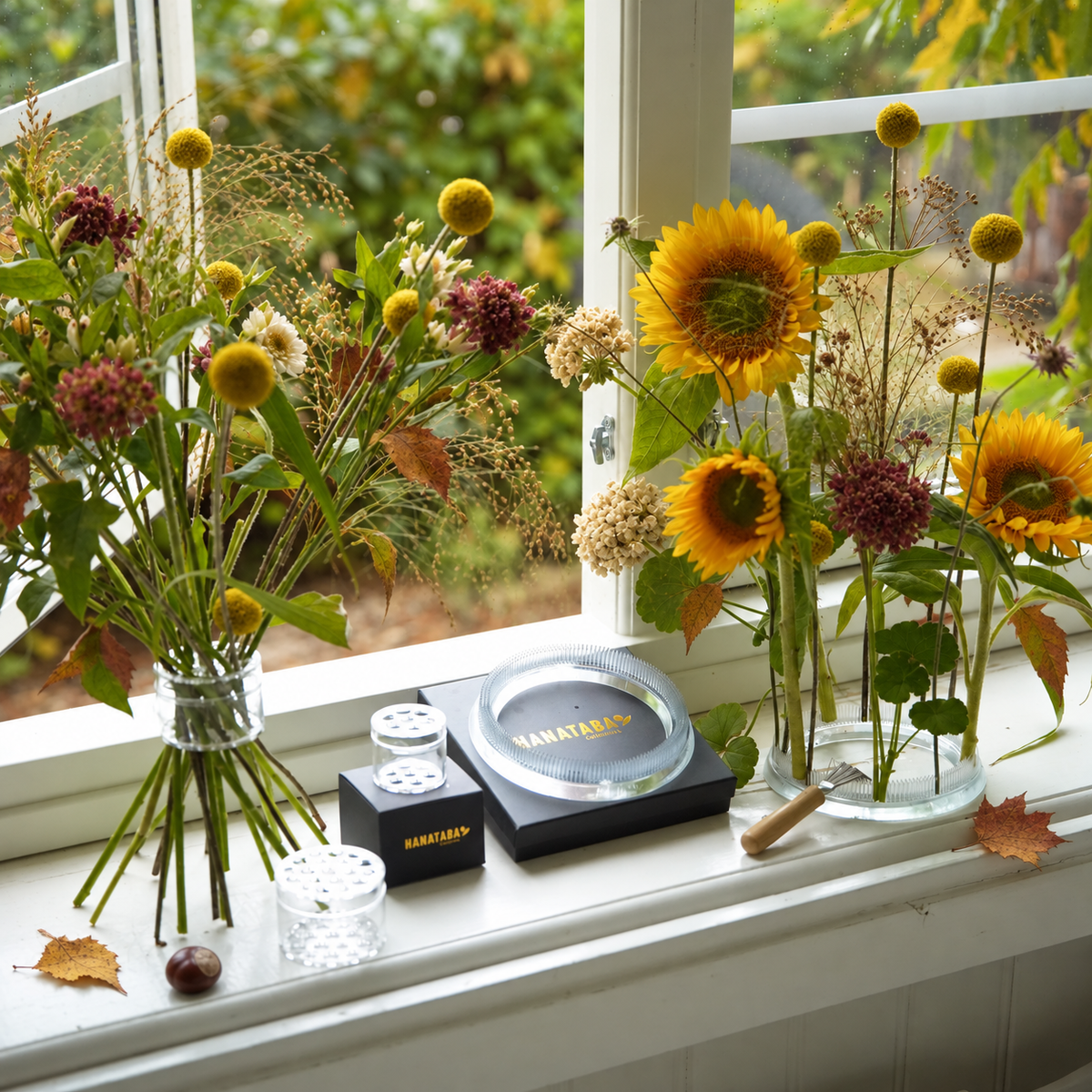 A styled arrangement of dried and fresh flowers on a white windowsill, featuring sunflowers, globe flowers, and wild grasses in glass vases, alongside a acrylic 'HANATABA' flower tools and accessories.