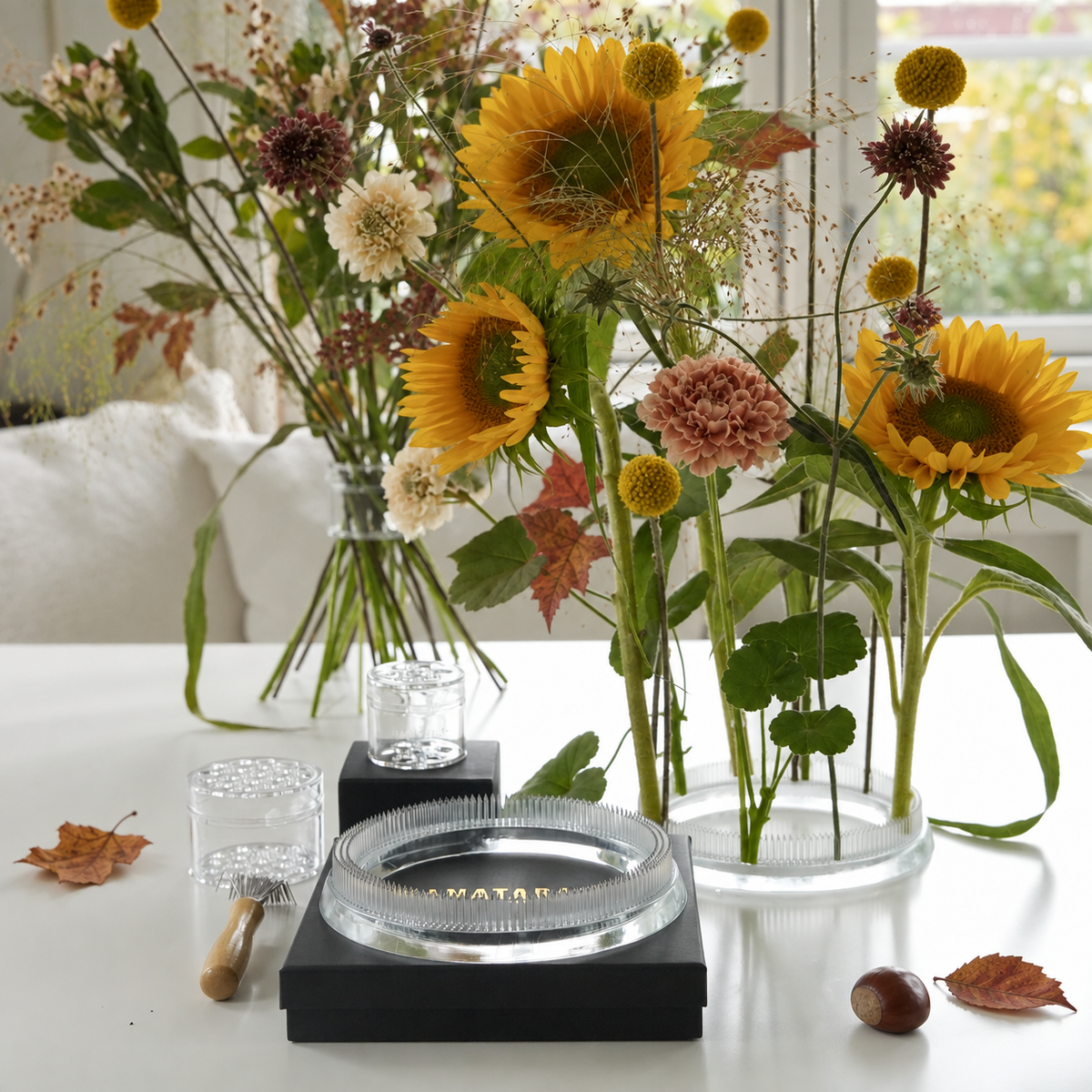 A styled tabletop arrangement with one hanataba bouquet twister crystal clear and one Hanataba Original Kenzan ring 150 mm acrylic, filled with sunflowers and dried flowers, set on a white surface with a black tray, Hanataba Original flower tools, and scattered autumn leaves in a bright indoor setting.