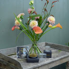 A floral arrangement in a Crystal Clear Hanataba Bouquet Twister displayed on a rustic wooden table, with flower tools including a kenzan 70 mm and textra silicon discs. The background is a painted green wooden wall.