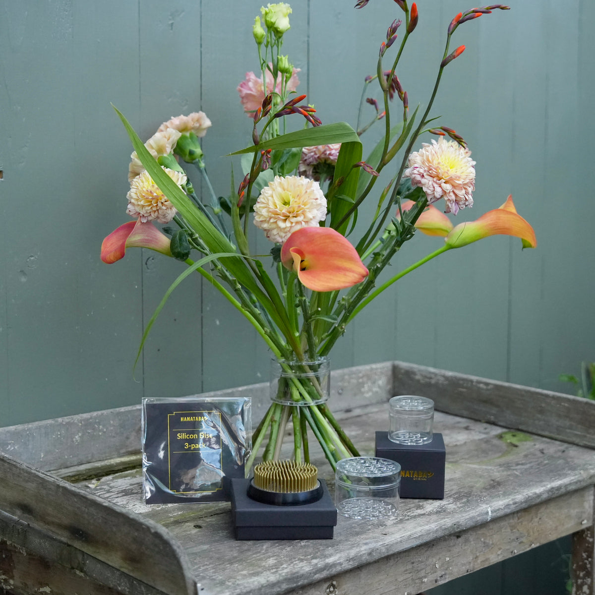 A floral arrangement in a Crystal Clear Hanataba Bouquet Twister displayed on a rustic wooden table, with flower tools including a kenzan 70 mm and textra silicon discs. The background is a painted green wooden wall.