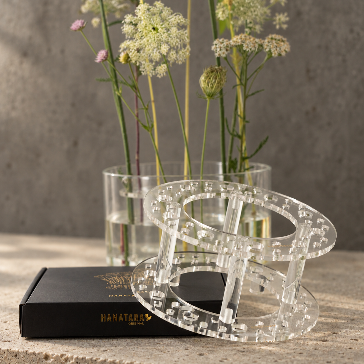 A clear acrylic two-tier stand with a geometric block pattern, displayed on a black Hanataba box with dried flowers in the background.