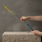 A person's hands holding a long flower stem over a textured stone block, using a bright blue curved tool to trim or cut the stem against a dark gray wall background.