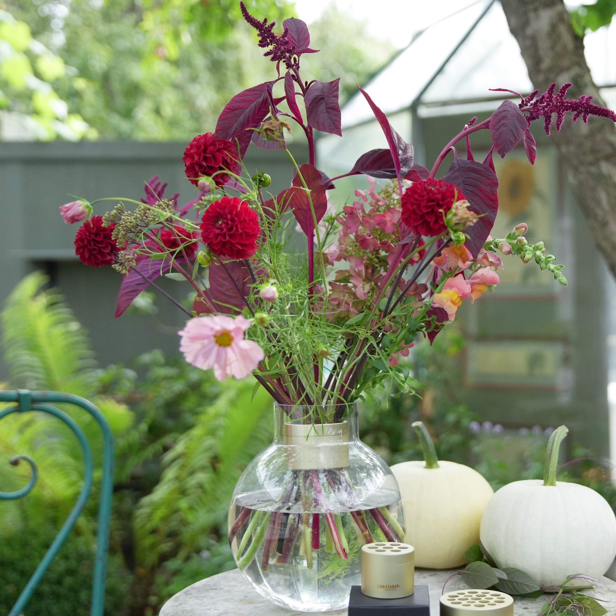 Hanataba Champagne Gold Bouquet Twister with red and pink flowers in a clear vase with pumpkins on a table outdoors.