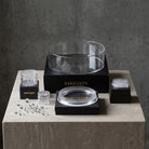 Hanataba clear glass and acrylic floral arrangement set arranged on a stone pedestal against a gray concrete wall, including a large clear bowl on a black base, smaller clear containers, a clear lid, and a white scrubber brush.