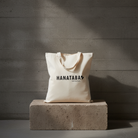 Beige tote bag with 'HANATABA' branding on a concrete block against a gray concrete wall.