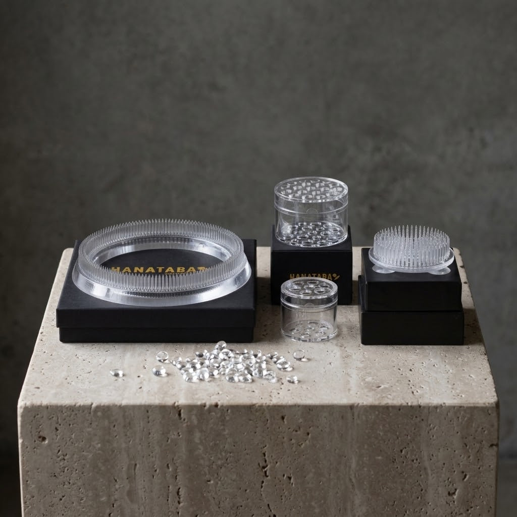 A set of clear acrylic flower arrangement tools, including a round kenzan ring 150 mm on a black and gold HANATABA branded box, two small cylindrical hanataba bouquet twister crystal clear, and a acrylic kenzan 70 mm, arranged on black boxes with scattered clear rhinestones on a concrete pedestal against a dark gray background.