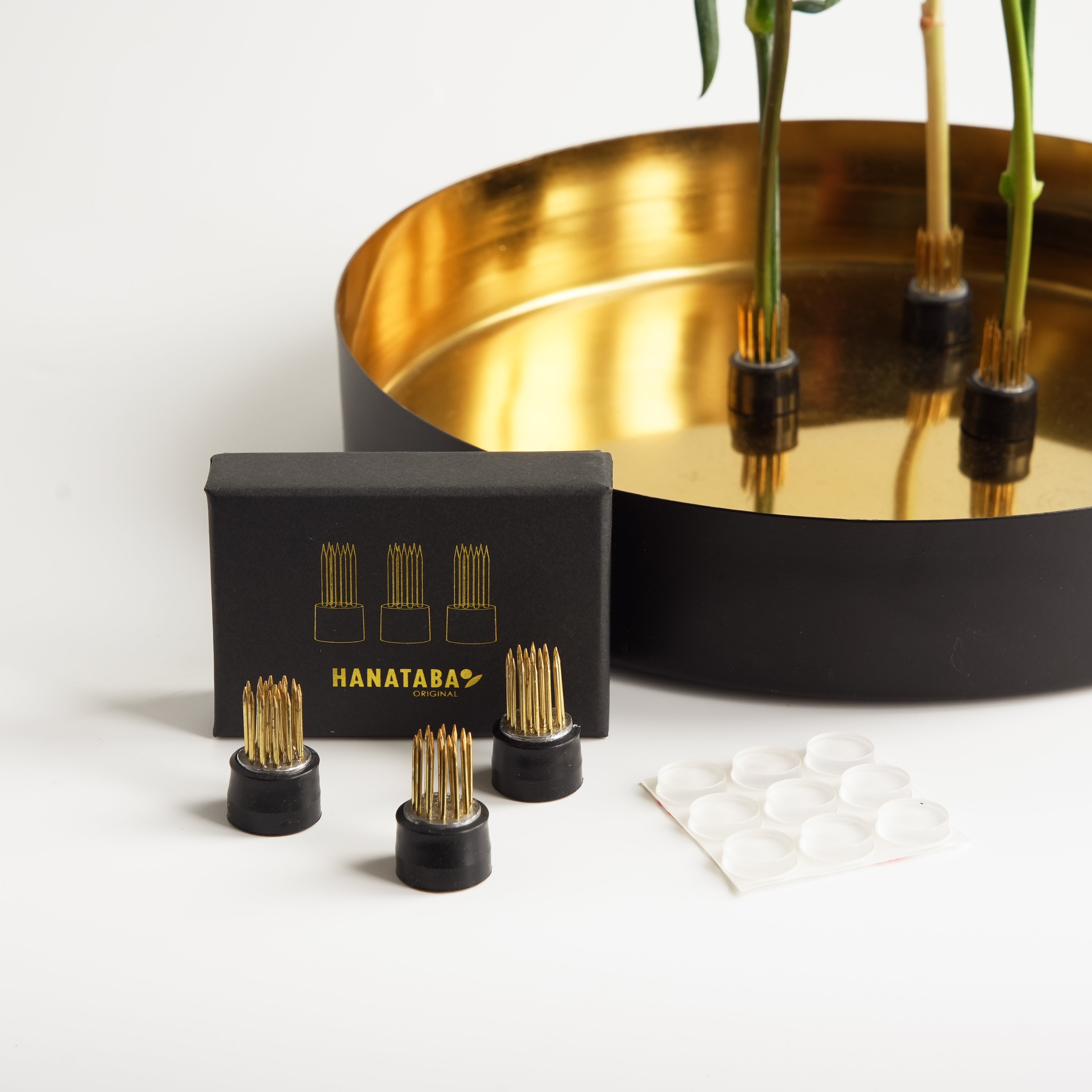 Hanataba magnetic flower frog set with three small gold pin frogs, magnets, and silicone pads displayed beside a gold-lined bowl holding flower stems.