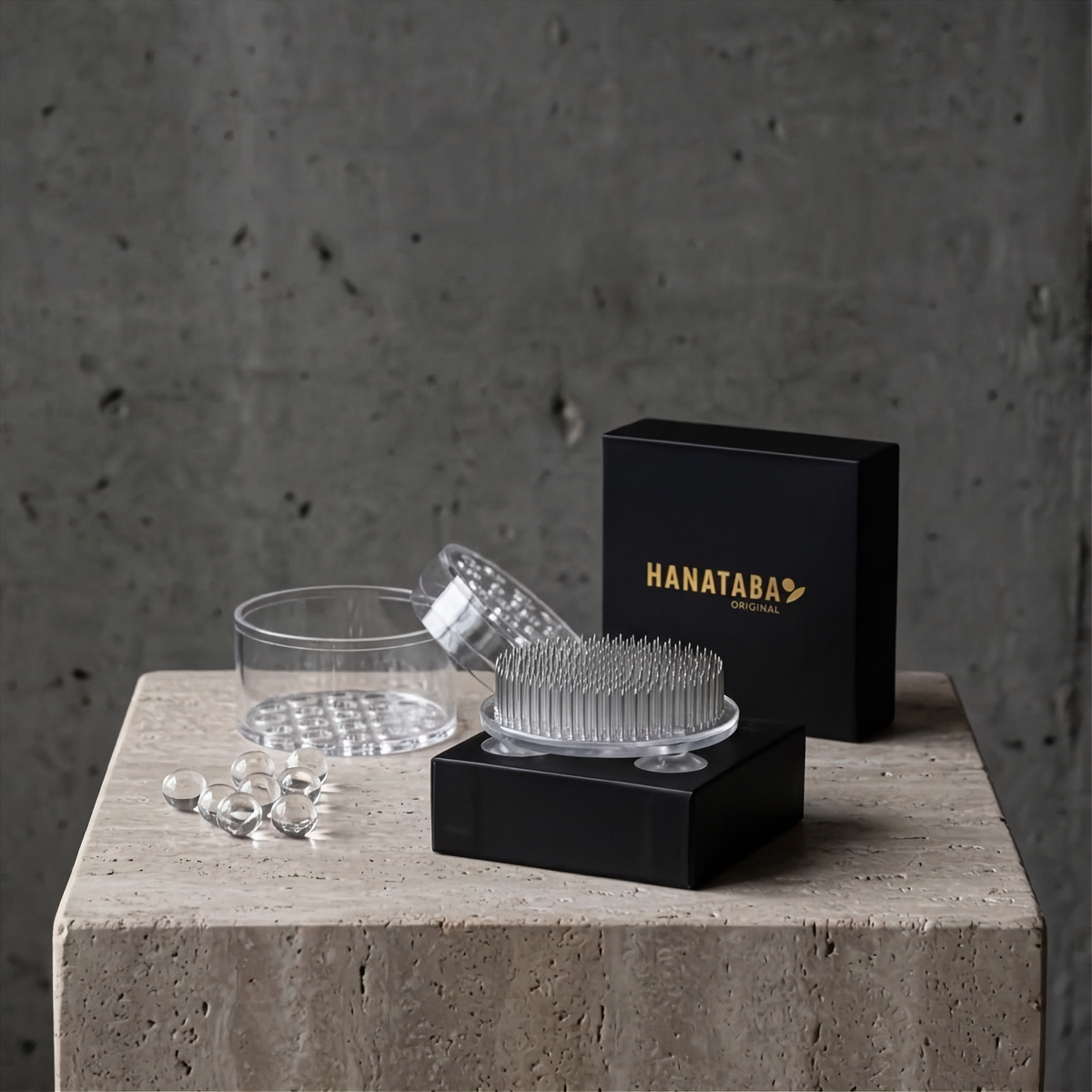A clear acrylic Hanataba transparent set displayed on a stone pedestal against a concrete wall, including transparent kenzan 70 mm, its black presentation box, a Hnataba Bouquet Twister Crystal Clear, and a few water beads.