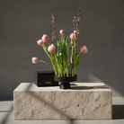 Black ceramic ikebana vase with flower arrangement including pink tulips and green leaves on a stone surface against a dark background.