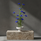 White ceramic ikebana pedestal vase with blue flowers on a stone block against a dark gray background