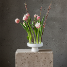 White vase with pink tulips and willow branches on a concrete block against a gray wall.
