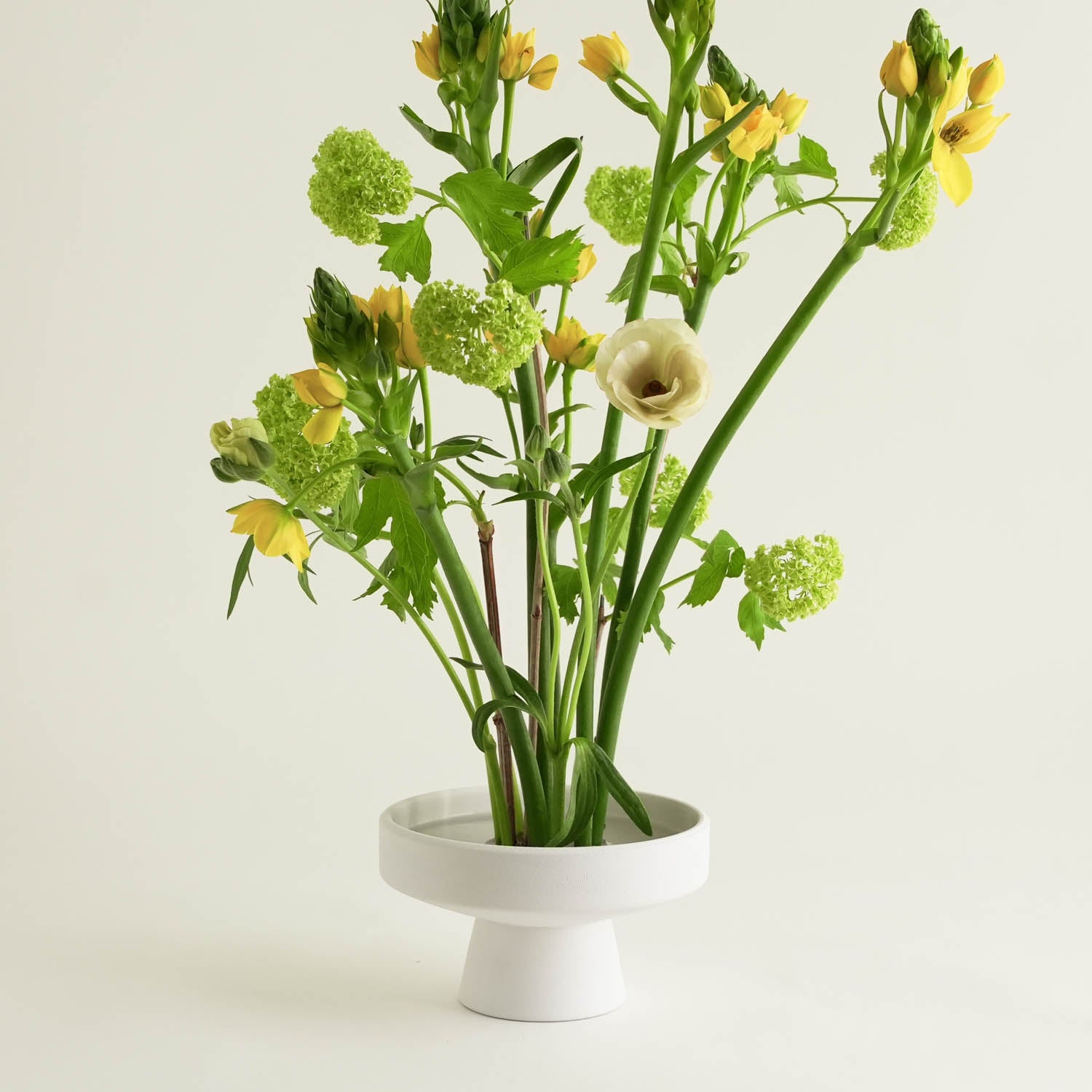 A white, pedestal-style vase with a Japanese ceramic touch holds an arrangement of yellow and white flowers alongside lush green foliage. Behind the vase is a black packaging box with the hanataba logo in gold, all set against a plain white background.