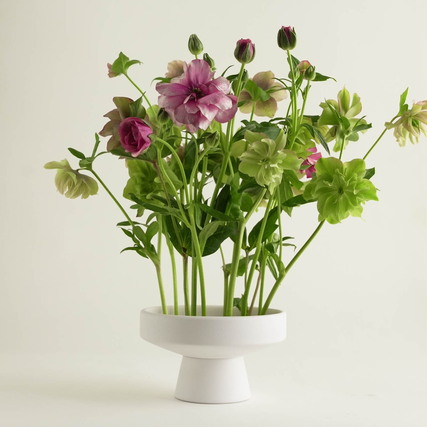 A white, pedestal-style vase with a Japanese ceramic touch holds an arrangement of pink flowers alongside lush green foliage. All set against a plain white background.