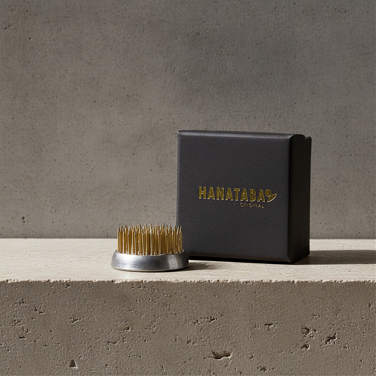 A Hanataba Original kenzan flower frog 34 mm with a silver metal base and gold-colored sharpened edges, displayed next to its black branded presentation box on a concrete ledge against a textured gray wall.