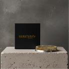 A Hanataba Original Kenzan Ring 120 mm with a circular base sits on a textured concrete pedestal next to a black square box printed with the brand name 'HANATABA ORIGINAL' in gold, set against a gray concrete wall.