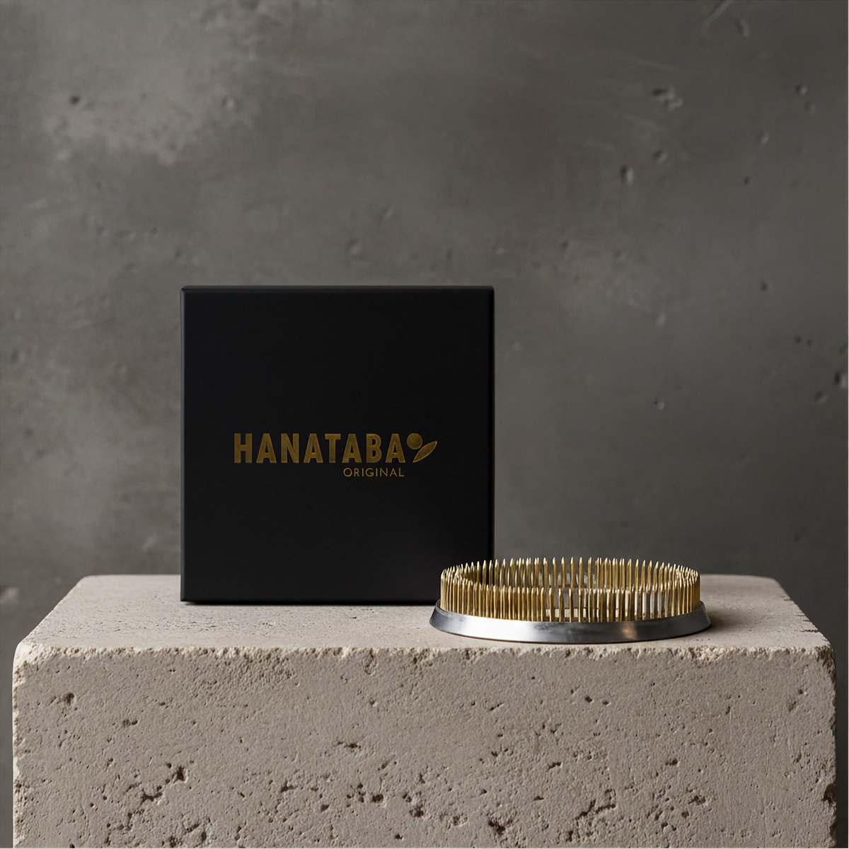 A Hanataba Original Kenzan Ring 120 mm with a circular base sits on a textured concrete pedestal next to a black square box printed with the brand name 'HANATABA ORIGINAL' in gold, set against a gray concrete wall.