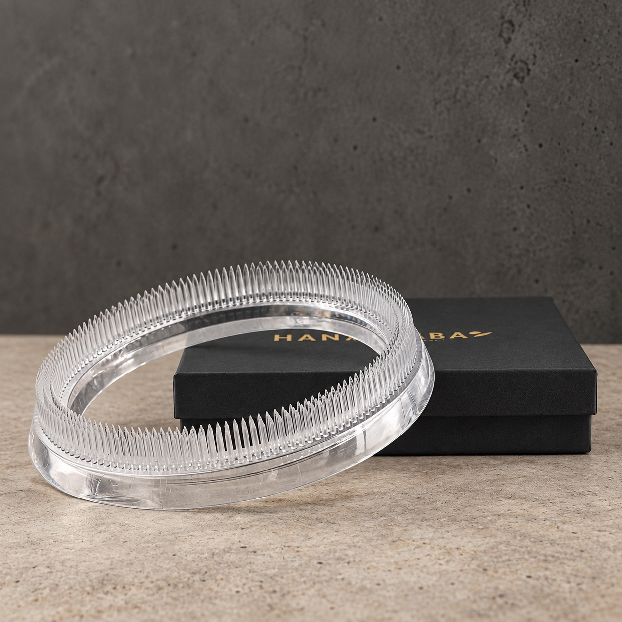 A clear acrylic ring-shaped kenzan flower frog 150 mm in acrylic material with fine bristles, shown in front of a black HANATABA box on a textured beige surface against a dark gray wall.
