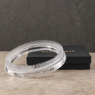 A clear acrylic ring-shaped kenzan flower frog 150 mm in acrylic material with fine bristles, shown in front of a black HANATABA box on a textured beige surface against a dark gray wall.