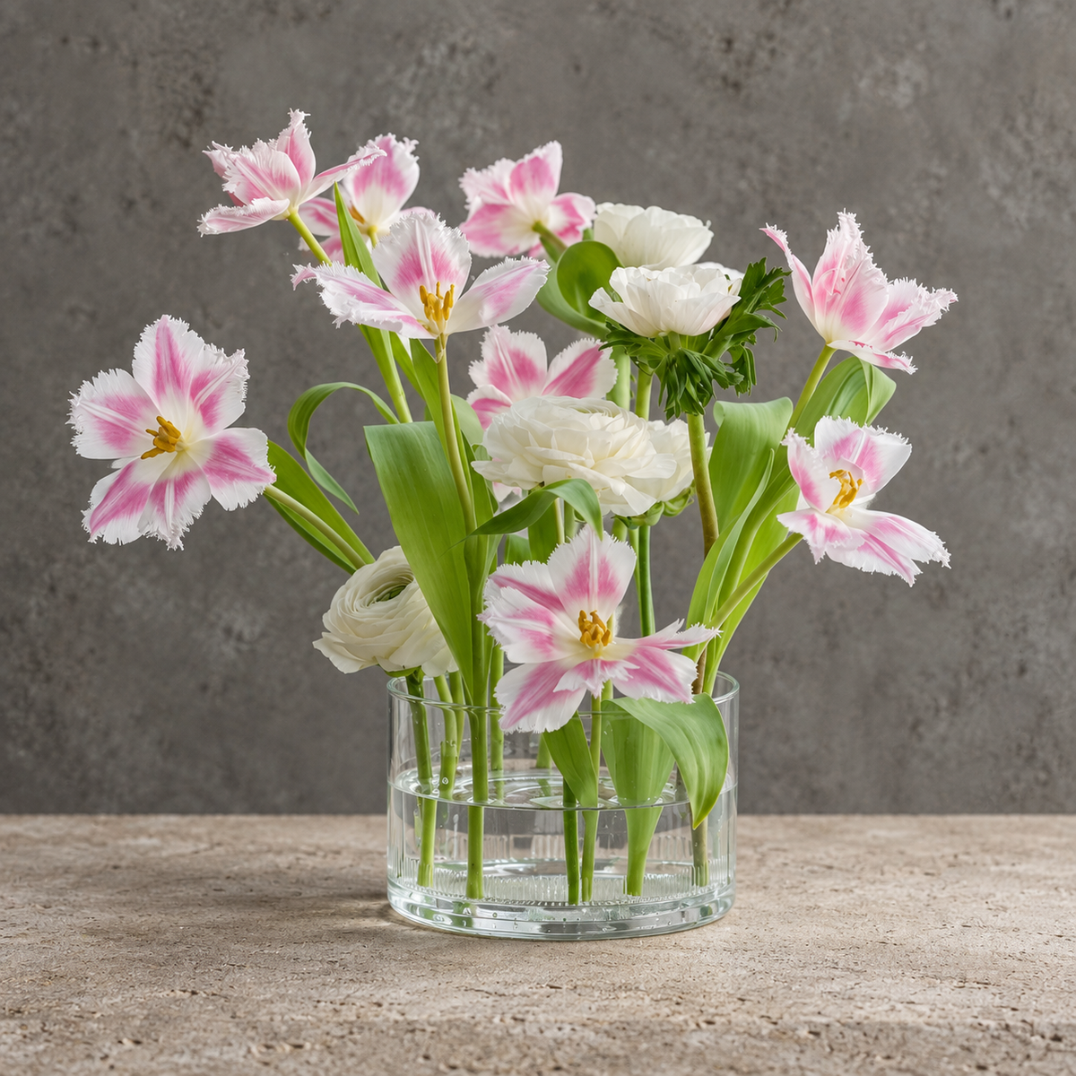 A flower arrangement with flowers in a clear glass vase, featuring pink and white tulips and white roses, set against a textured gray background.