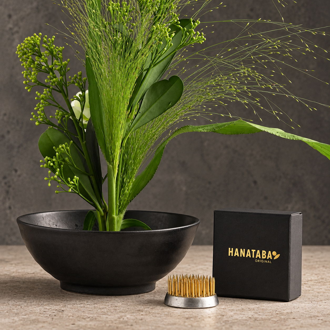 A Hanataba kenzan flower frog 34 mm with brass bristles and a silver metal base, displayed next to its black presentation box on a stone surface. In the background, a black ceramic bowl holds a green floral arrangement against a textured gray wall.