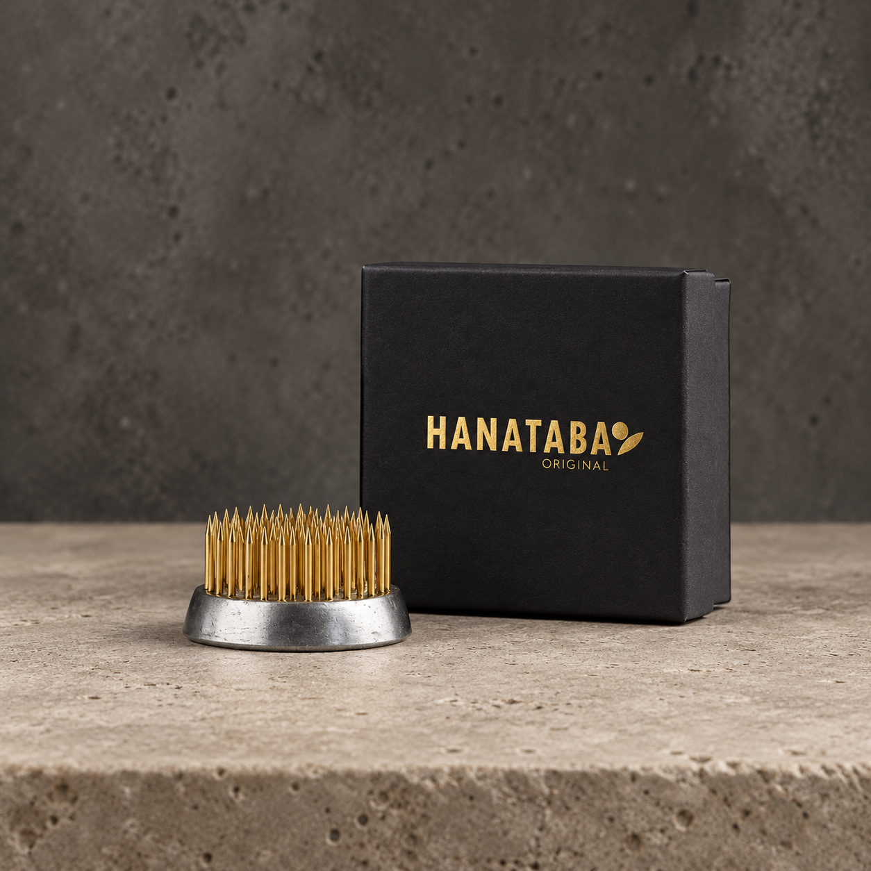 A Hanataba Original kenzan 34 mm with gold-colored bristles and a silver metal base, displayed next to its black branded box on a textured stone surface.